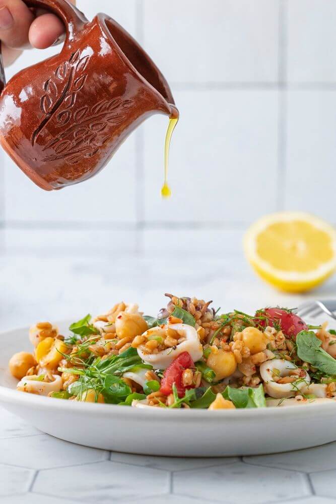 Hand holding a small brown ceramic pitcher, pouring golden olive oil over a colorful chickpea and vegetable salad on a white plate, with a halved lemon blurred in the background.