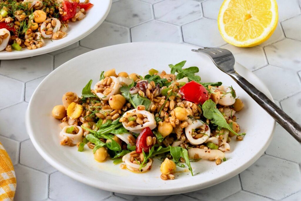 White plate filled with a fresh chickpea and vegetable salad featuring arugula, cherry tomatoes, grains, and squid rings, accompanied by a fork and a halved lemon on a hexagonal tiled surface.