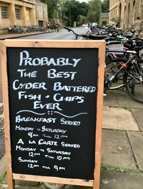 Wooden framed chalkboard sign on a sidewalk advertising "Probably The Best Cyder Battered Fish & Chips Ever," with breakfast and a la carte serving hours listed, positioned near a row of parked bicycles on a street with buildings and trees in the background.