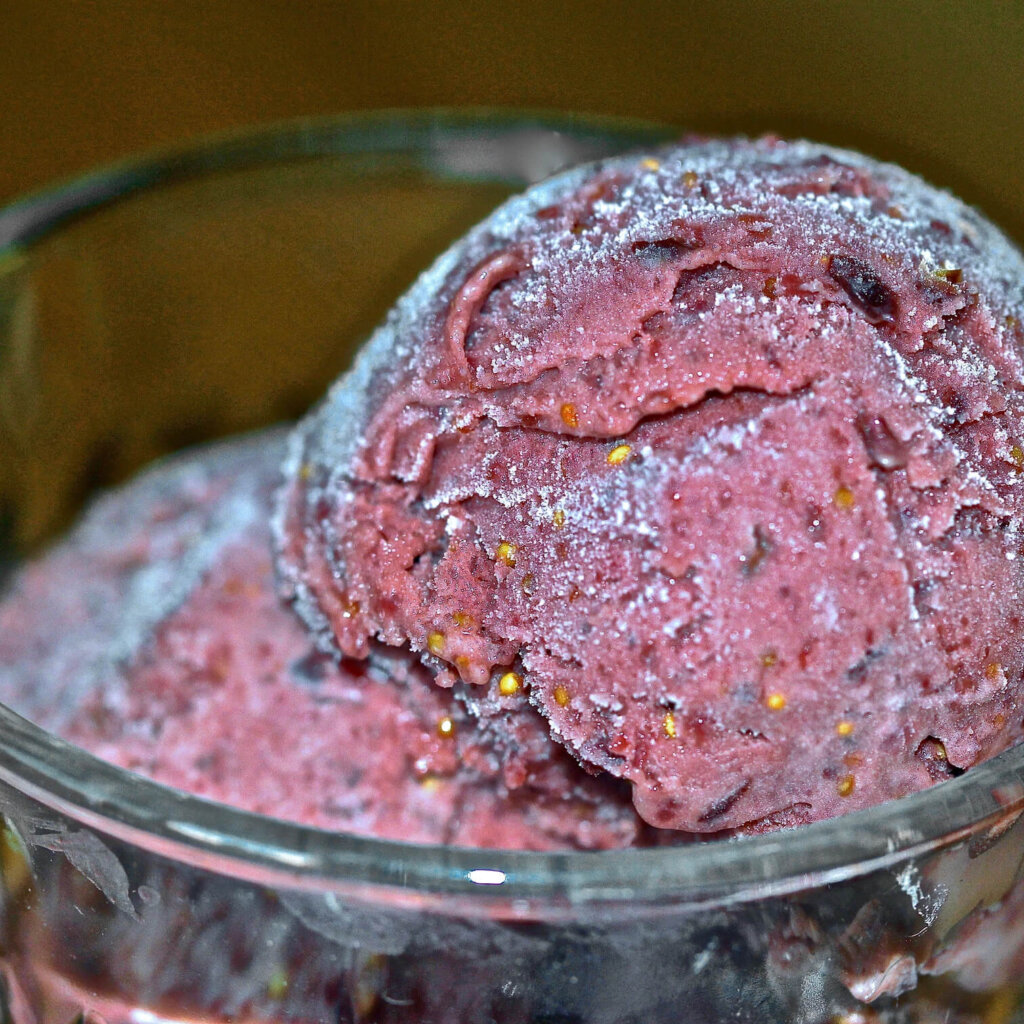 Close-up image of two scoops of fig ice cream in a clear glass bowl, showing the creamy texture and visible fig seeds sprinkled throughout the rich purple-pink ice cream.