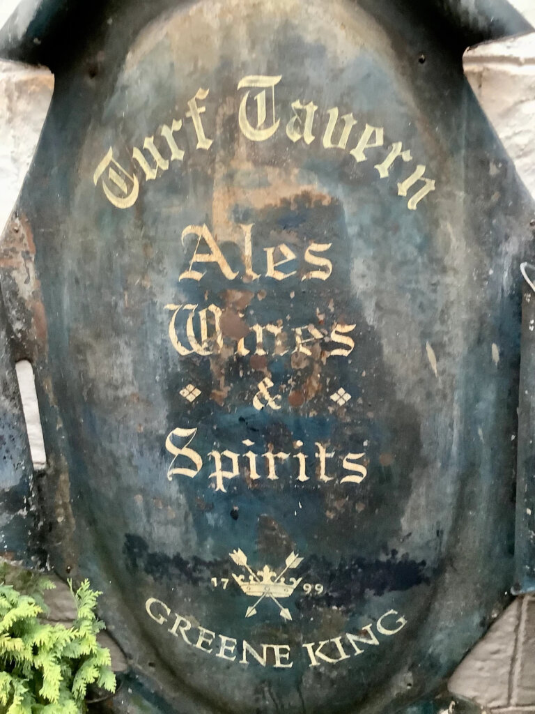 A weathered metal pub sign with gold lettering reading "Turf Tavern Ales Wines & Spirits 1799 Greene King," featuring a crown and crossed arrows emblem near the bottom.