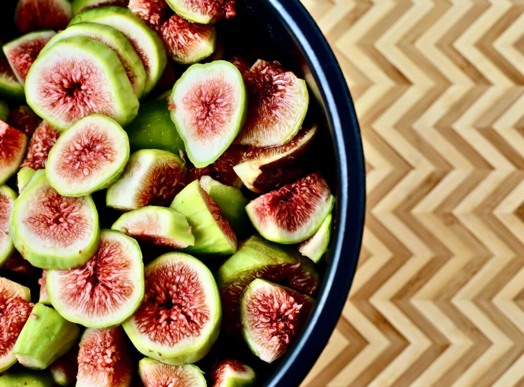 Top-down view of a black bowl filled with freshly sliced green figs, showcasing the pinkish-red interior and seeds, placed on a wooden surface with a chevron pattern.